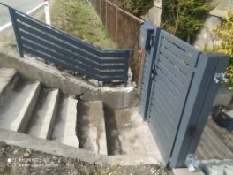 Nowoczesna, grafitowa furtka z poziomymi szczeblami, zamontowana przy betonowych schodach i ogrodzeniu z siatki. Ujęcie z dołu, widok na otwartą furtkę.