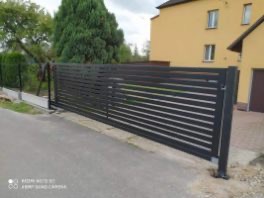 Nowoczesna, przesuwna brama wjazdowa z poziomych, grafitowych profili, na tle zieleni i żółtego domu. Widoczne detale konstrukcyjne i mechanizm bramy.