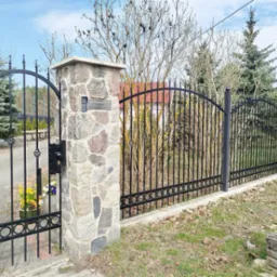 Elegancka, czarna brama z kutymi zdobieniami, osadzona na kamiennych słupkach, z widocznym domofonem i kwiatami w donicy. Tło stanowią drzewa i błękitne niebo.