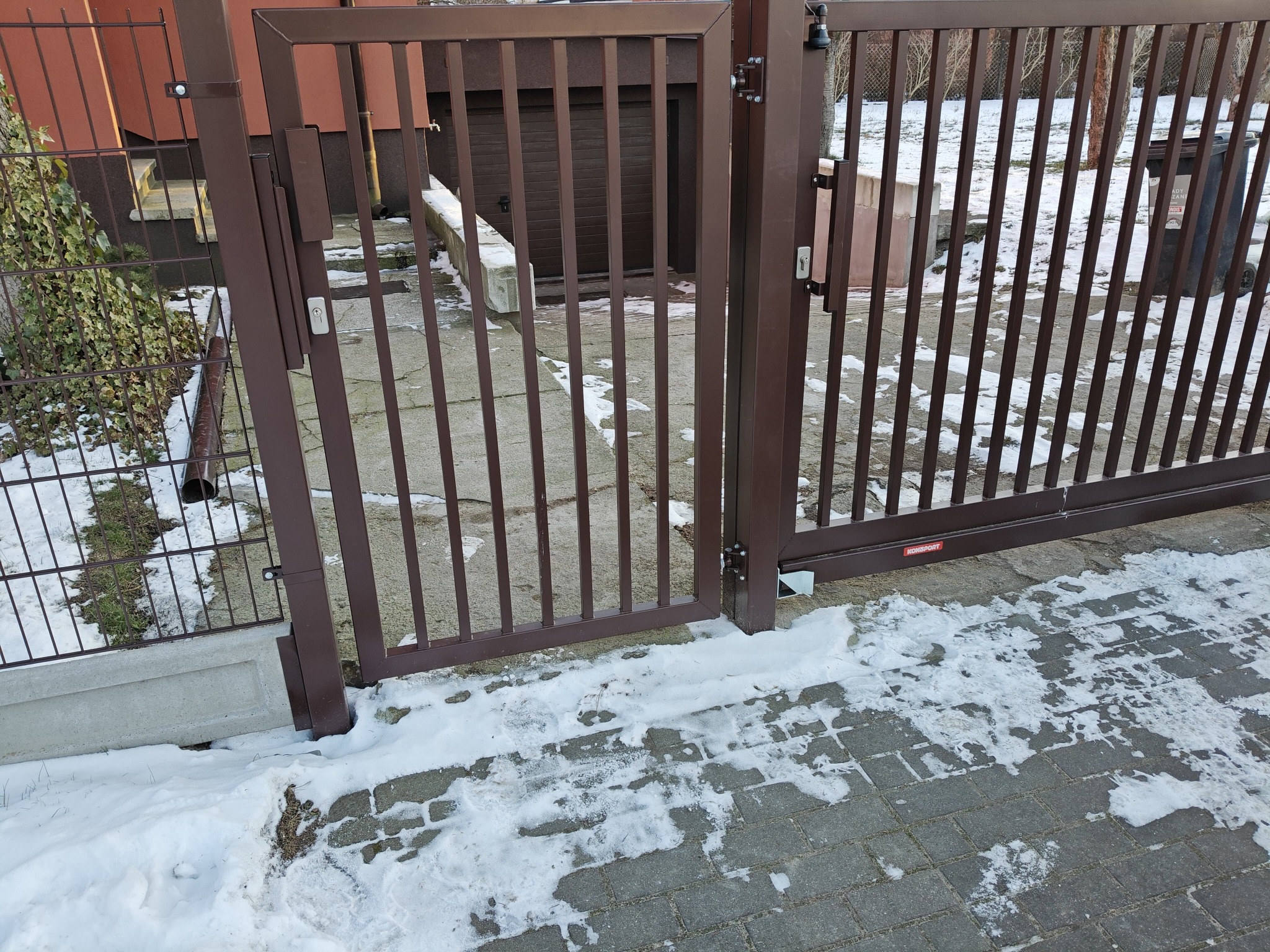 Otwarta, brązowa brama metalowa z pionowymi szczeblami na posesji pokrytej śniegiem. Widoczny fragment ogrodzenia panelowego i brama garażowa w tle.