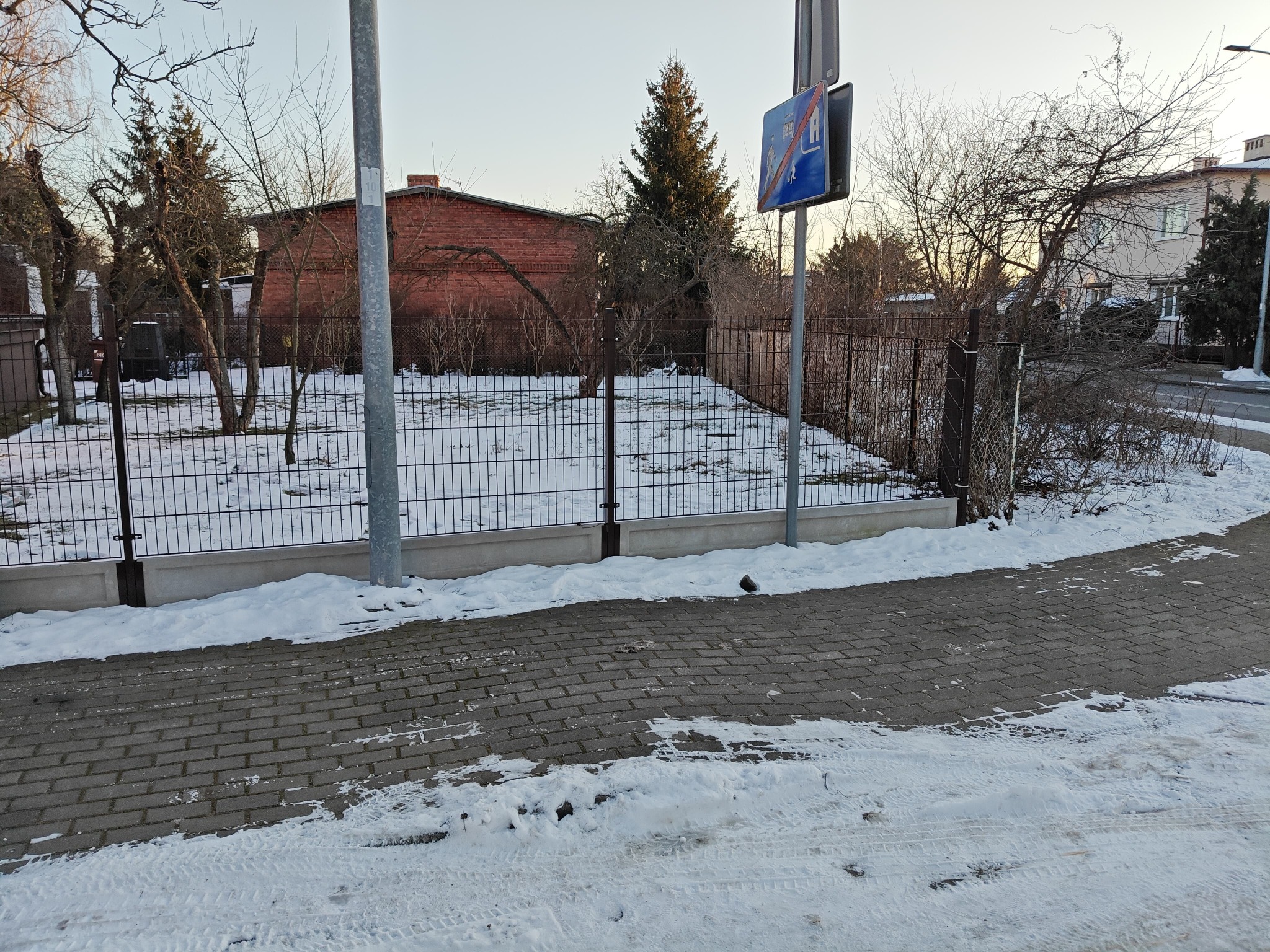 Zimowy krajobraz z czarnym ogrodzeniem panelowym na betonowej podmurówce, widoczne z chodnika. W tle budynek z czerwonej cegły i drzewa.