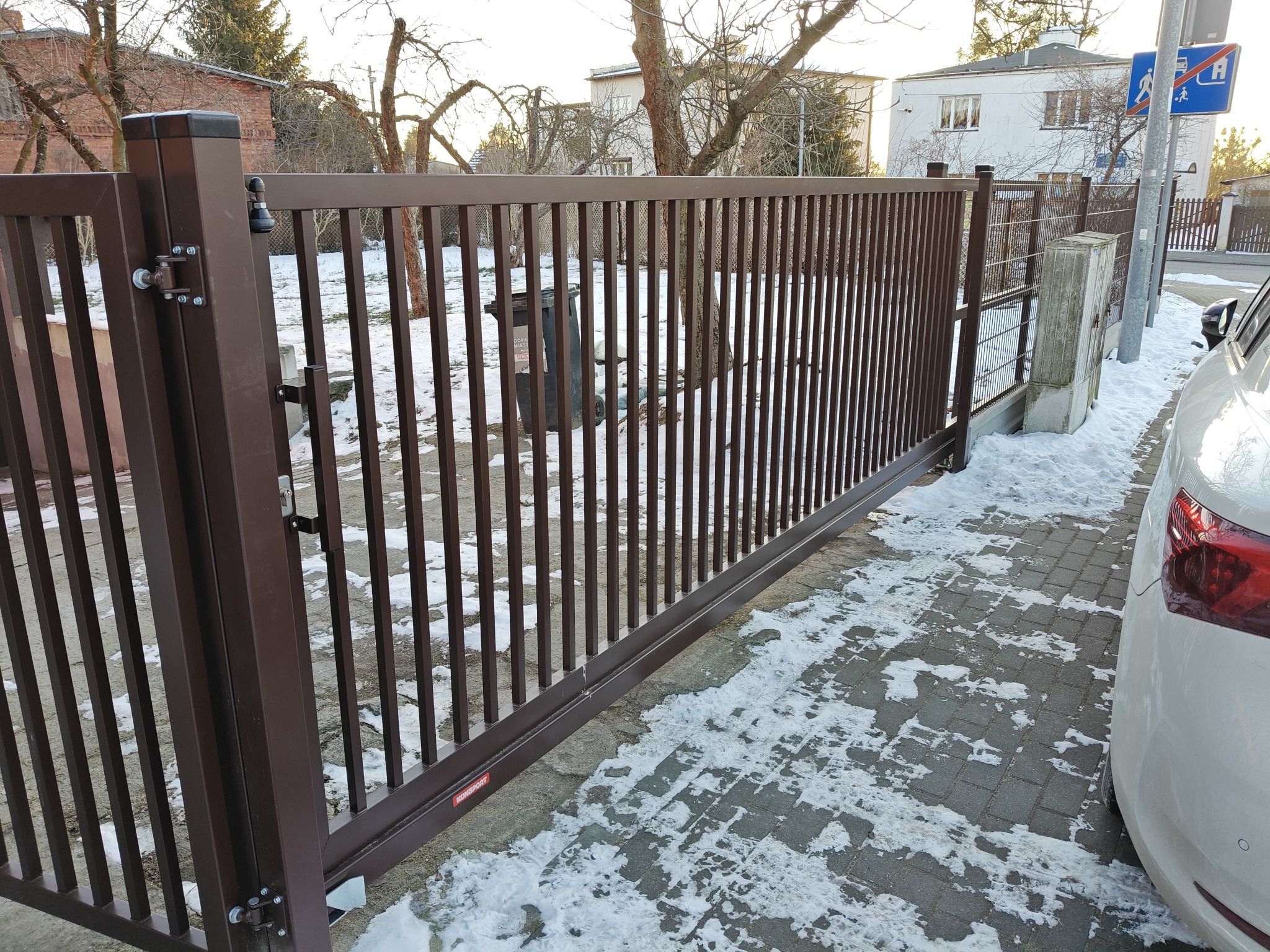 Nowoczesna, przesuwna brama metalowa w kolorze brązowym na tle zimowego krajobrazu. Widoczny fragment samochodu i kostka brukowa z resztkami śniegu.