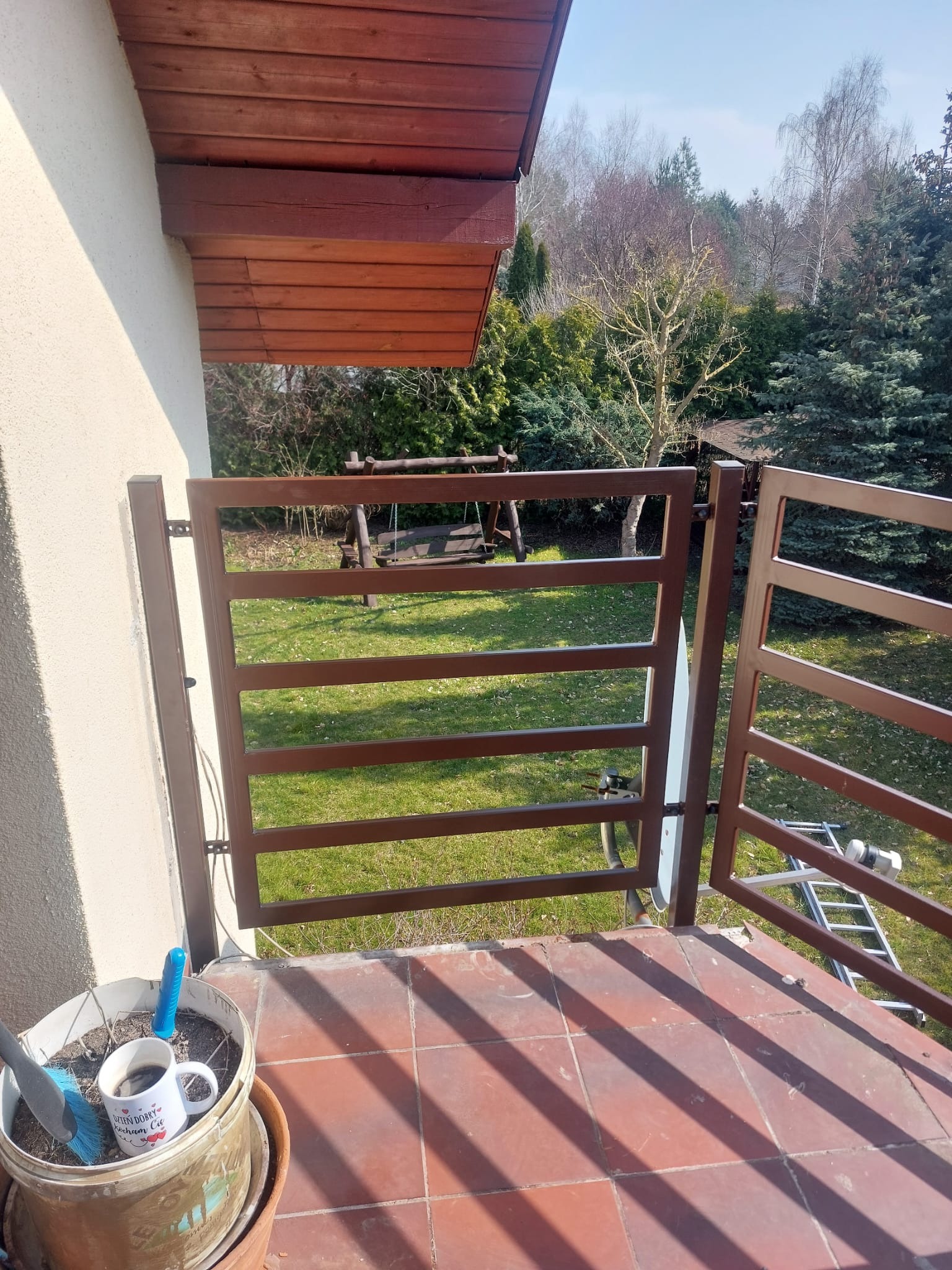 Balkon z brązową, metalową balustradą o nowoczesnym designie. Na pierwszym planie donica z kwiatami i kubek. W tle trawnik i drzewa. Widok z góry.