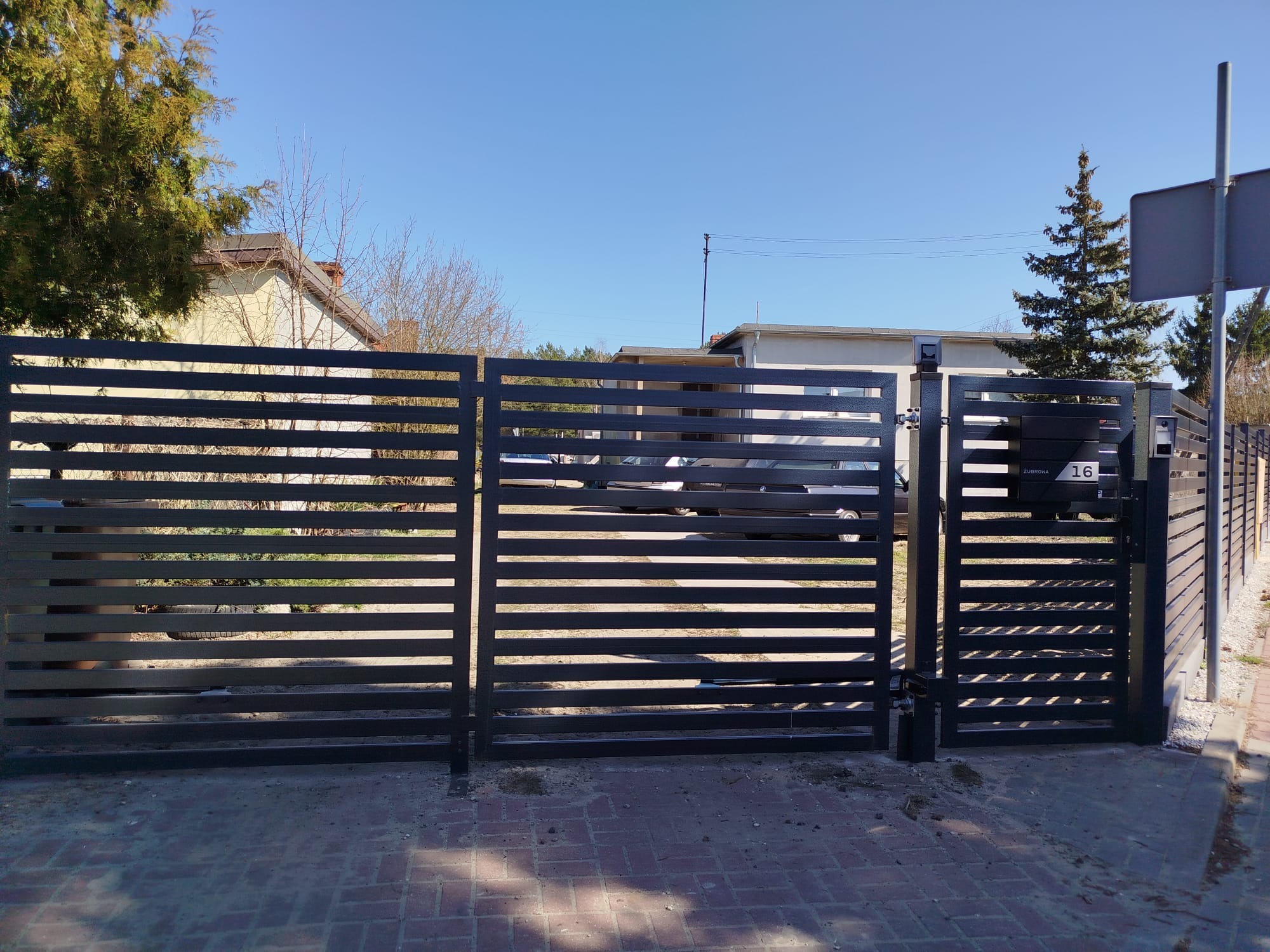 Nowoczesne, poziome ogrodzenie metalowe w kolorze antracytowym z bramą uchylną, numer posesji 16, widok z zewnątrz na tle zabudowy jednorodzinnej.