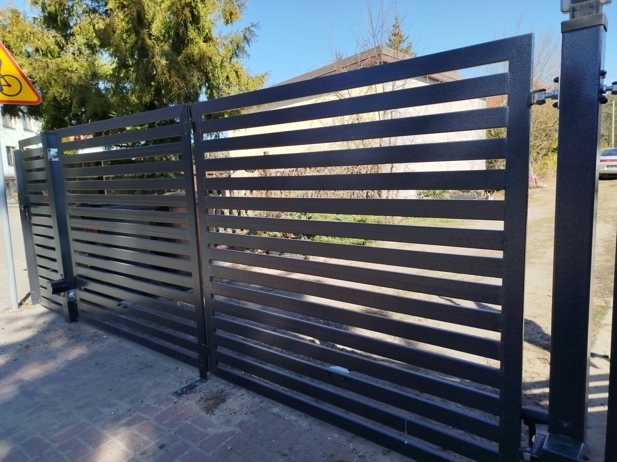 Nowoczesna, metalowa brama przesuwna z poziomymi szczeblami, pomalowana na ciemny kolor, zamontowana na posesji. Widoczny znak drogowy.