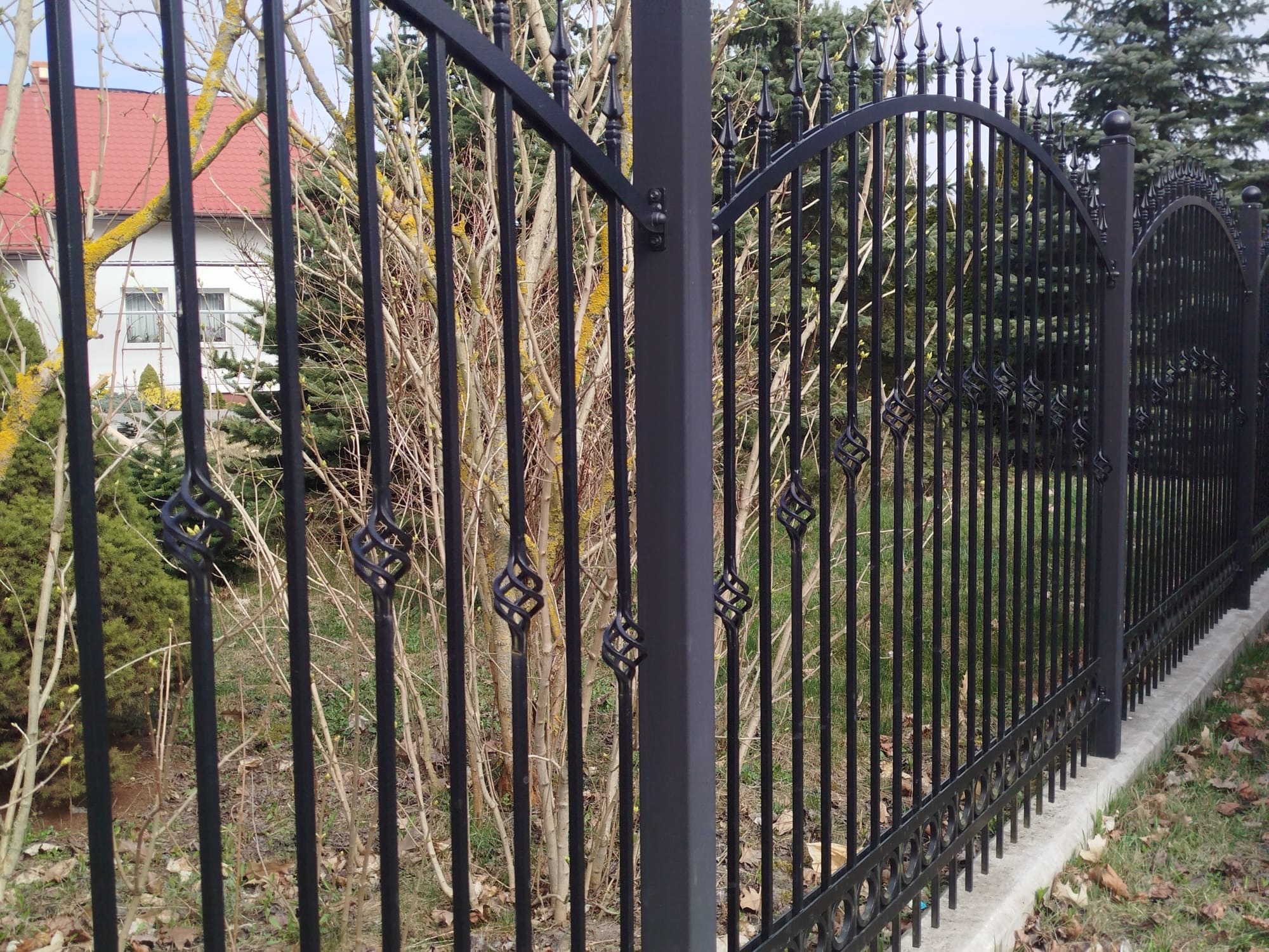 Czarne, kute ogrodzenie metalowe z ozdobnymi elementami w kształcie spirali na tle zieleni i domu z czerwoną dachówką. Widoczne słupki i podmurówka.