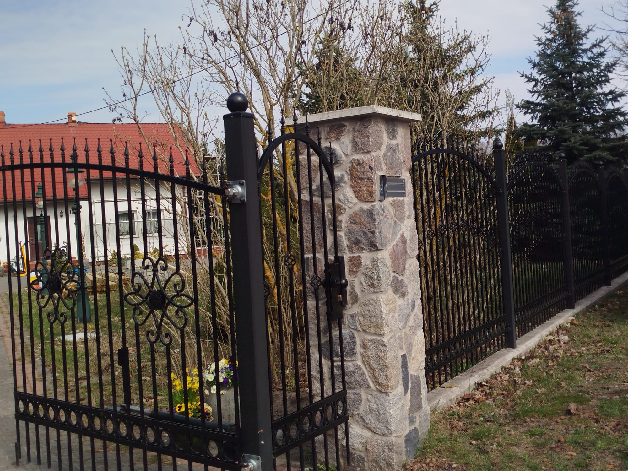Czarna, metalowa brama z dekoracyjnymi elementami i słupkiem z kamienia. W tle dom z czerwoną dachówką i zieleń.
