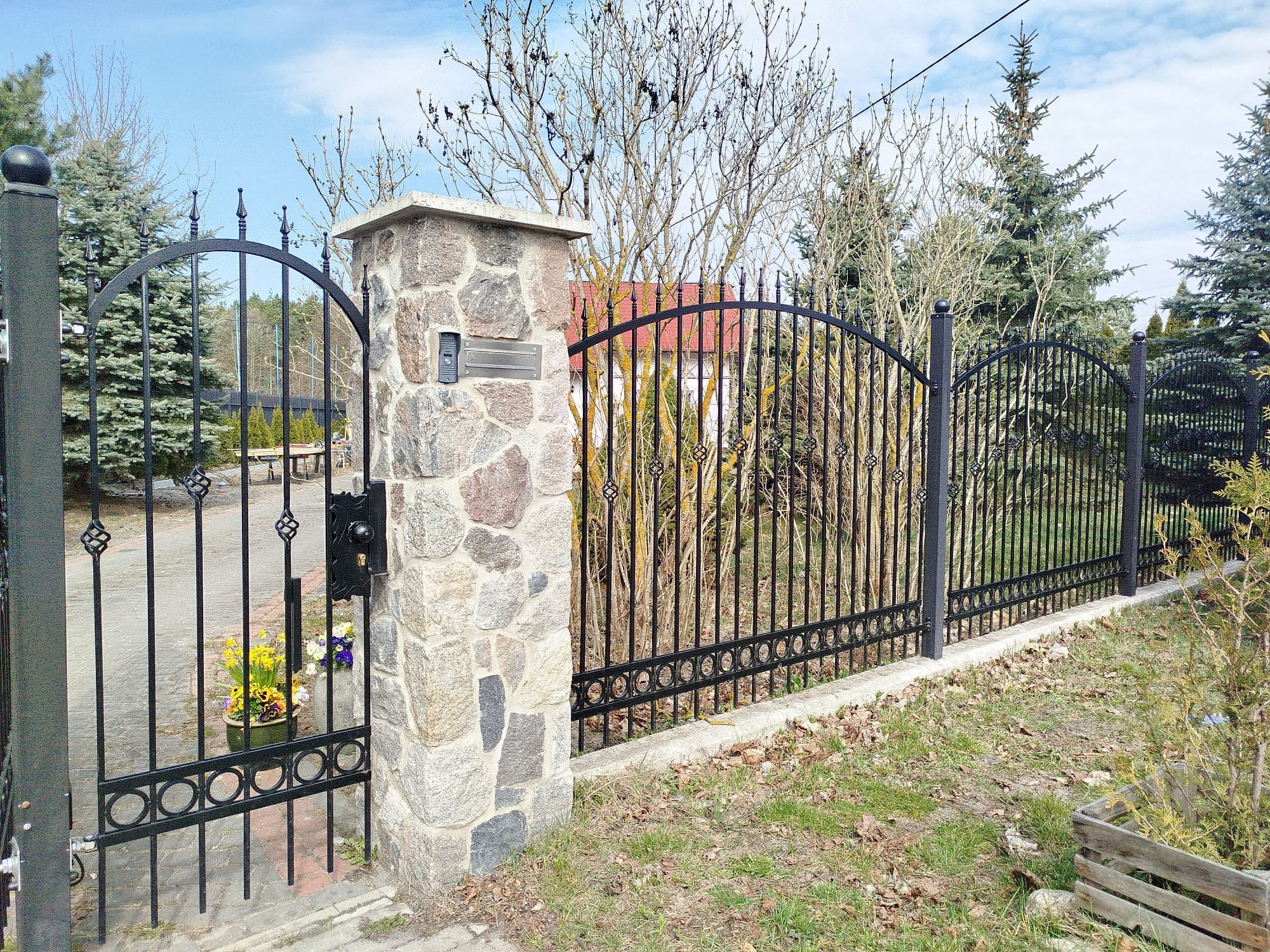 Elegancka, czarna brama z kutymi zdobieniami, osadzona na kamiennych słupkach, z widocznym domofonem i kwiatami w donicy. Tło stanowią drzewa i błękitne niebo.
