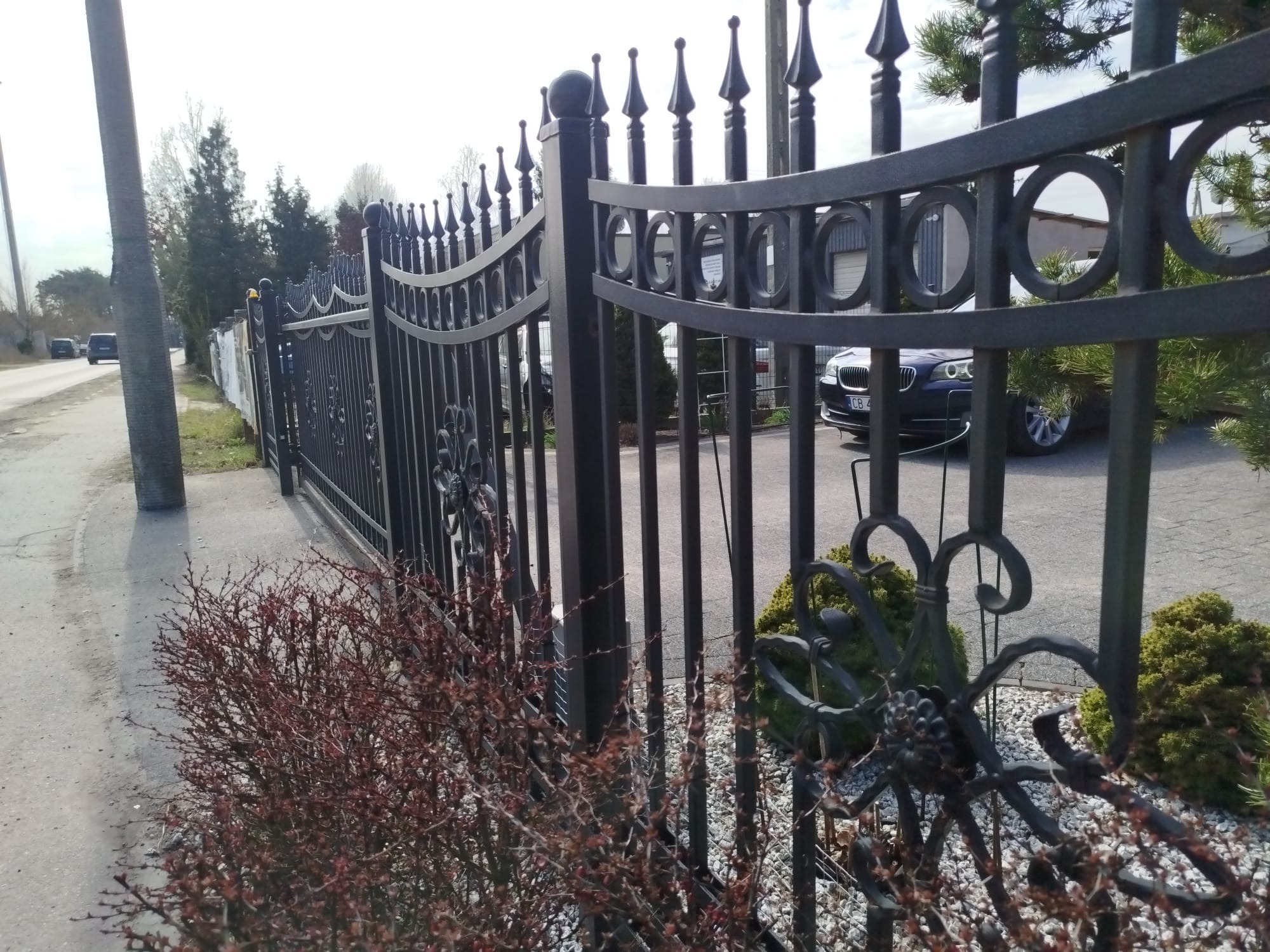 Eleganckie, kute ogrodzenie w kolorze grafitowym, z dekoracyjnymi elementami roślinnymi i geometrycznymi, oddziela posesję od ulicy. W tle widoczny samochód.