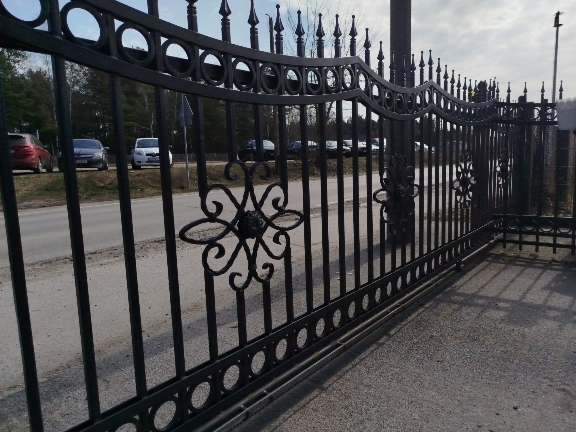 Elegancka, czarna brama przesuwna z metalu, zdobiona motywami roślinnymi i okręgami. Widok na ulicę i zaparkowane samochody w tle. Perspektywa z dołu.