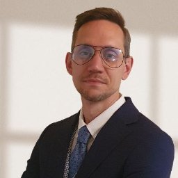 Łukasz Chojnowski Consulting - Mężczyzna w granatowym garniturze, białej koszuli i niebieskim, wzorzystym krawacie, w okularach aviatorach, z krótkimi brązowymi włosami i lekkim zarostem. Neutralne, jasne tło.