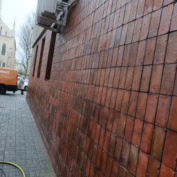 Clean -ART - Ceglana elewacja budynku po umyciu, widoczne mokre płytki i czysta powierzchnia. W tle fragment chodnika i budynek z wieżą. Wąż ciśnieniowy na chodniku.