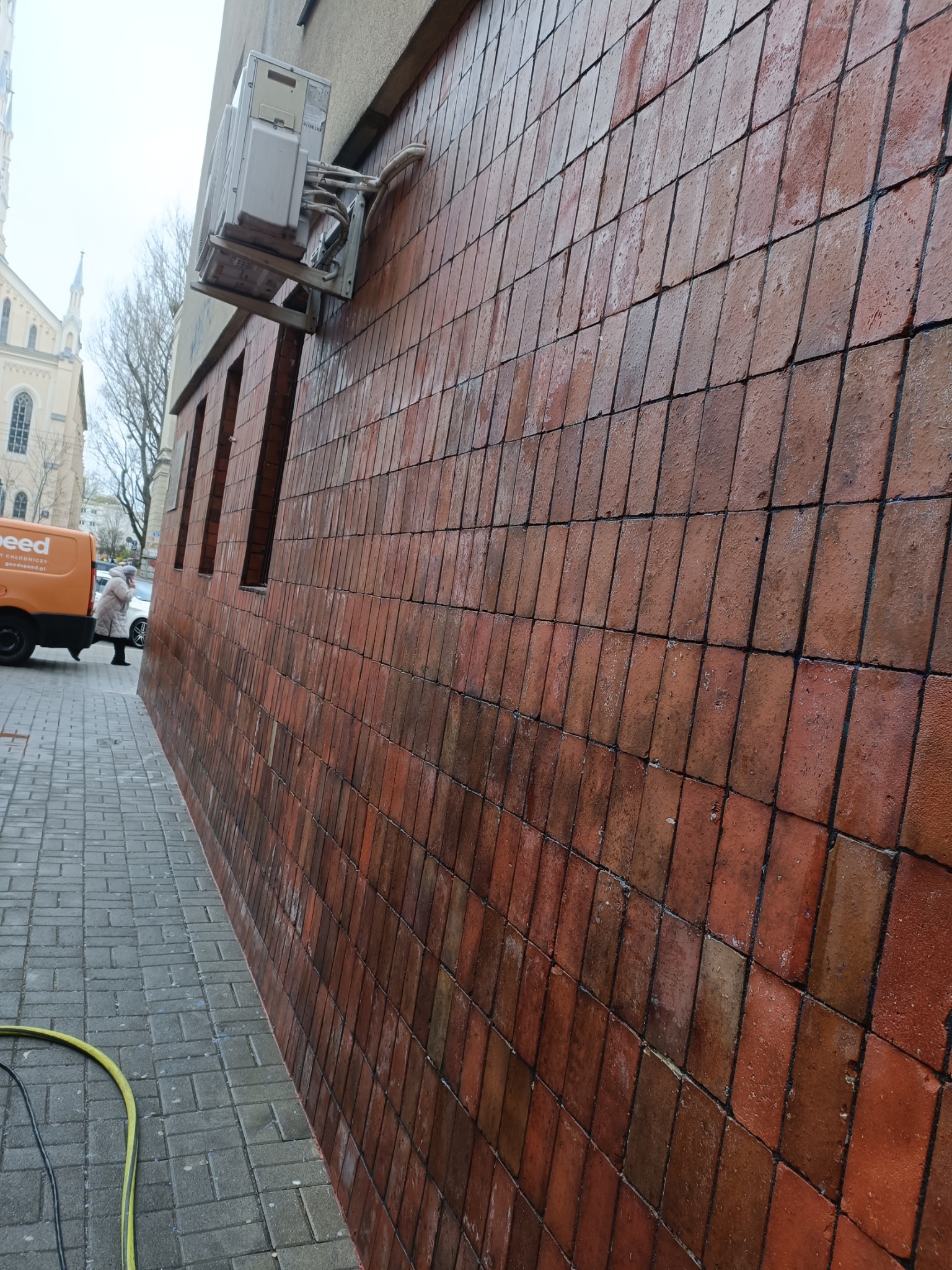 Ceglana elewacja budynku po umyciu, widoczne mokre płytki i czysta powierzchnia. W tle fragment chodnika i budynek z wieżą. Wąż ciśnieniowy na chodniku.