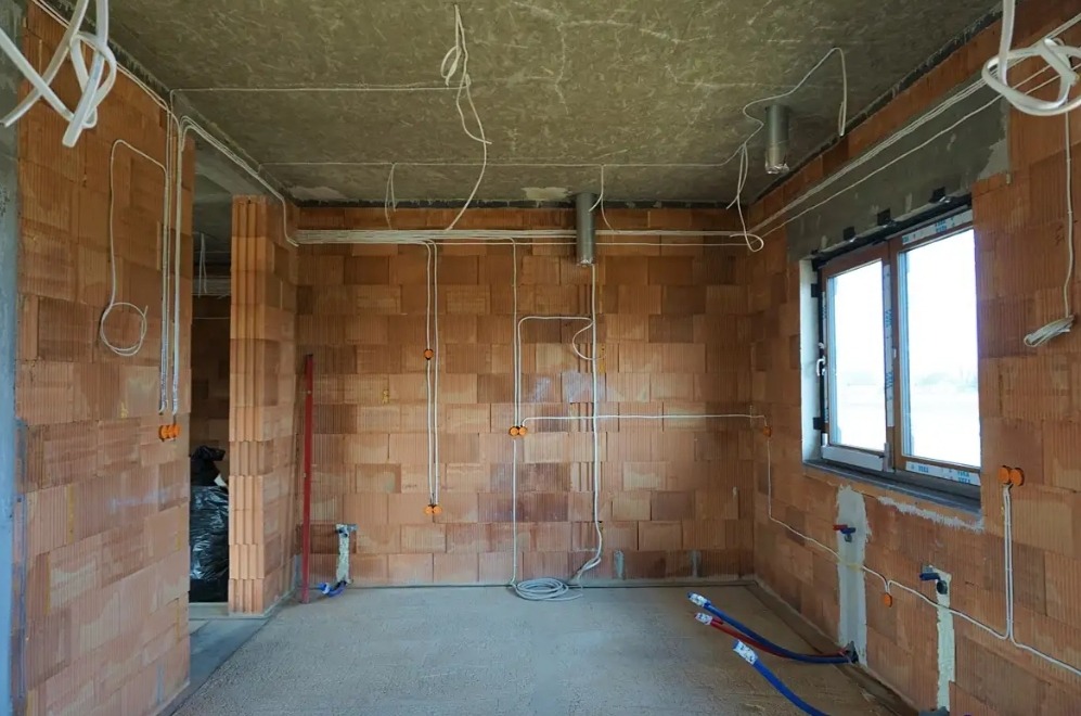 Wnętrze w trakcie prac: widoczne przewody elektryczne rozprowadzone po ścianach z cegły, puszki instalacyjne i fragment instalacji wodnej przy oknie.