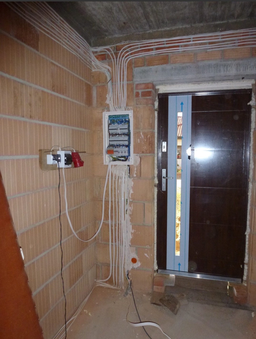 Rozdzielnica elektryczna z licznymi przewodami w nowym budynku z cegły, z gniazdkami i wtyczkami, obok drzwi wejściowych z niebieską folią ochronną.