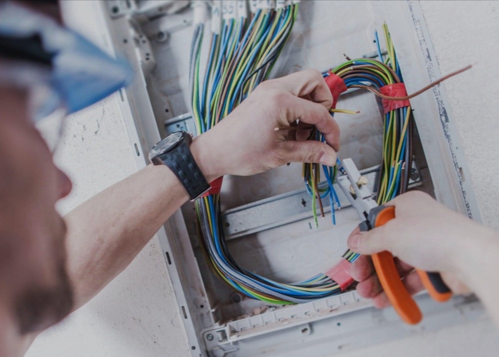 Elektryk w trakcie pracy: przycina przewody w skrzynce bezpiecznikowej, trzymając kombinerki z pomarańczową rączką. Widoczne kolorowe kable i zegarek na ręce.