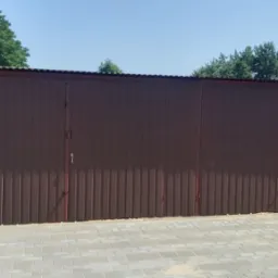 Brązowy garaż blaszany z dachem z blachy falistej na tle zieleni i nieba. Widoczne drzwi zamykane na klucz. Konstrukcja ustawiona na szarej kostce brukowej.