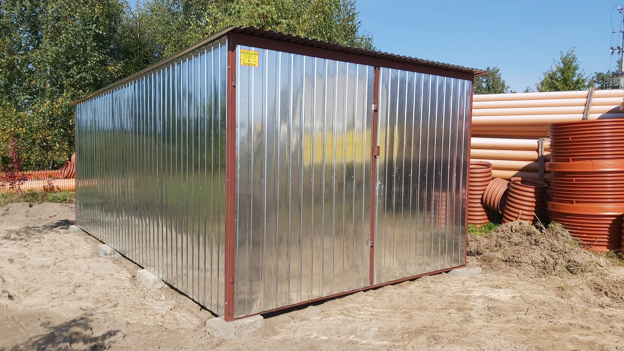 Błyszczący, metalowy garaż blaszany z brązowymi detalami na betonowych blokach, z widoczną naklejką informacyjną, otoczony ziemią i rurami.