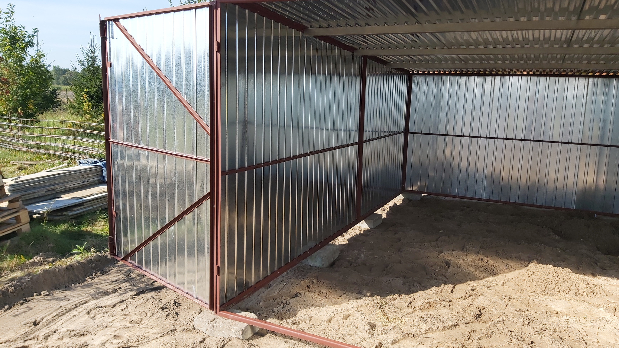 Metalowa konstrukcja garażu blaszanego z brązowymi profilami, widok z zewnątrz na piaszczyste podłoże wewnątrz. W tle drzewa i ułożone elementy budowlane.