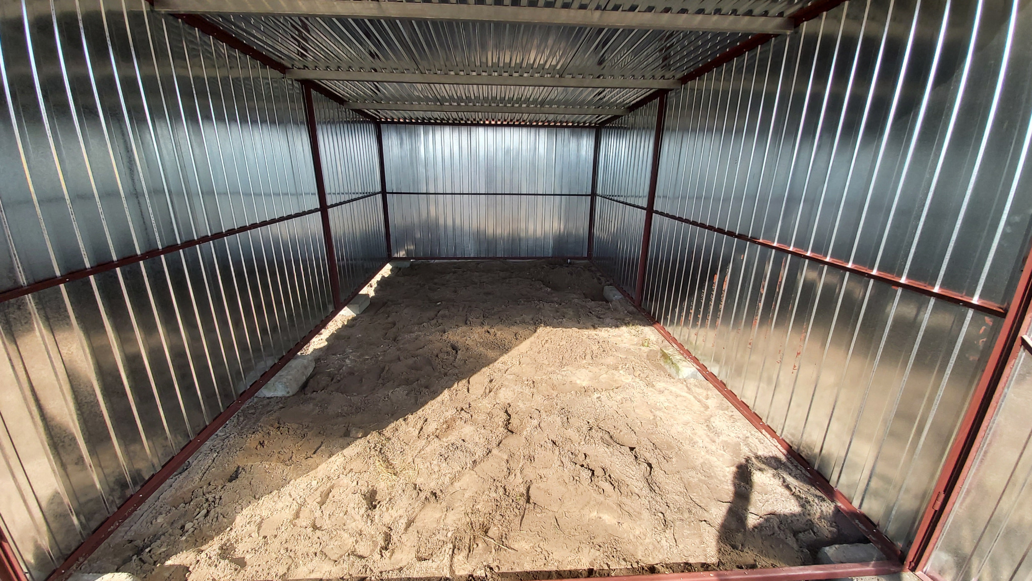 Wnętrze garażu blaszanego w Prażmowie z piaskiem na podłożu. Konstrukcja z metalowych profili i blachy falistej. Widok z przodu, wnętrze oświetlone.