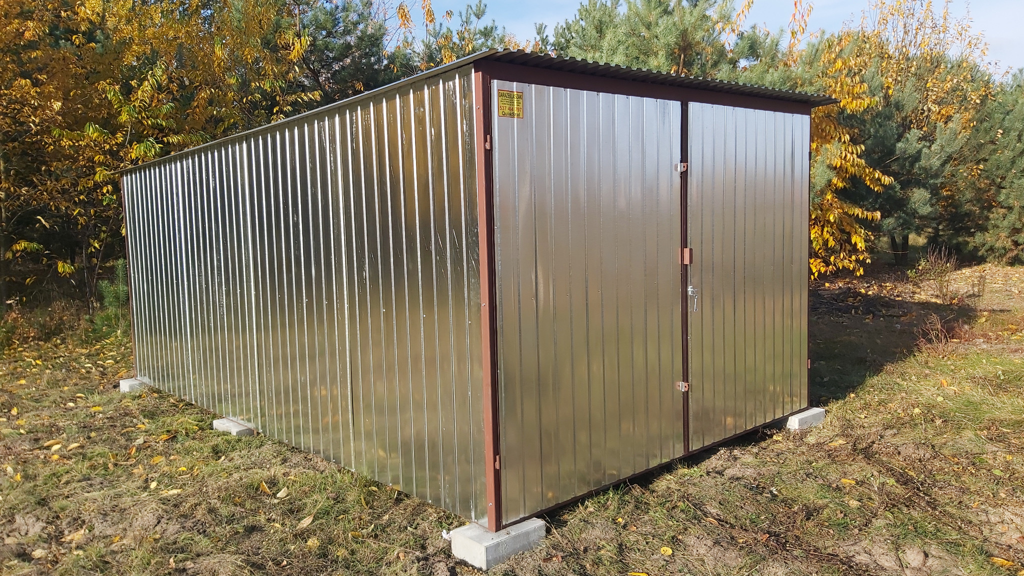 Zmontowany garaż blaszany z dwuskrzydłowymi drzwiami na betonowych bloczkach, otoczony jesienną zielenią. Widoczna naklejka producenta.