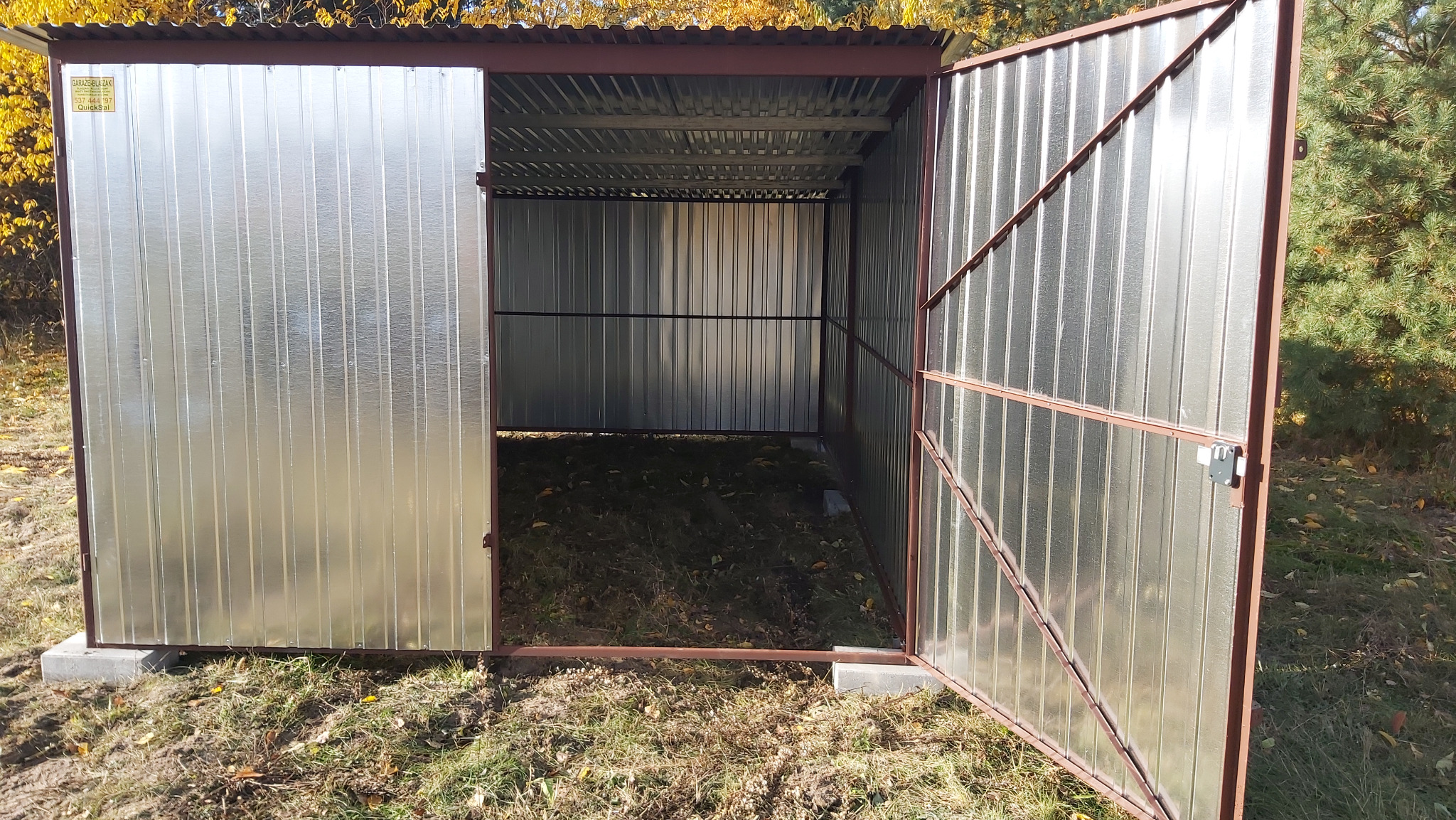 Otwarty, blaszany garaż z brązowymi ramami stoi na betonowych fundamentach na trawie. Widoczne jesienne liście i drzewa w tle. Metalowa konstrukcja, oświetlenie naturalne.