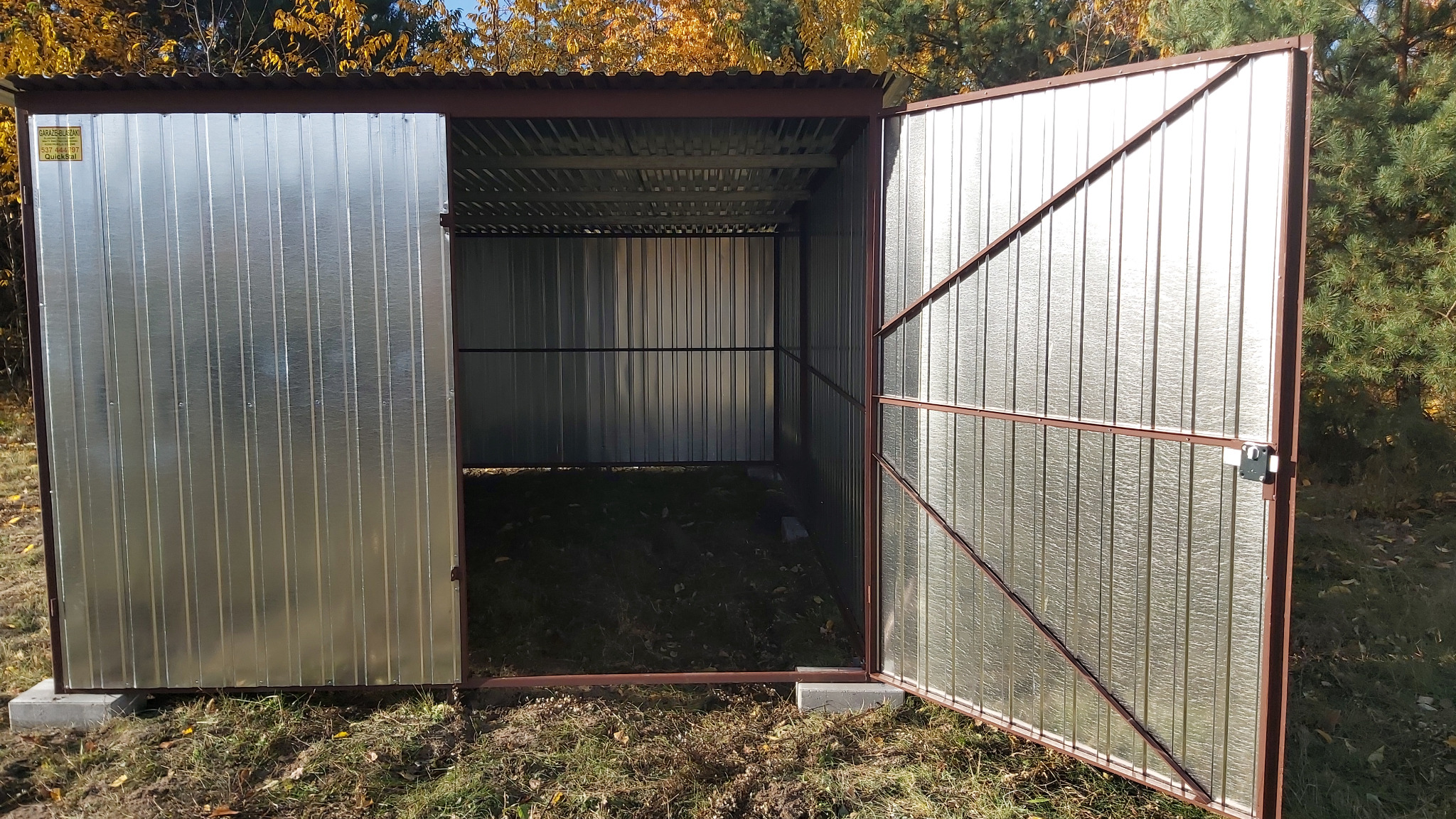 Otwarty, blaszany garaż z brązowymi obramowaniami na betonowych fundamentach, otoczony zielenią. Widoczna faktura blachy i jesienne drzewa w tle.