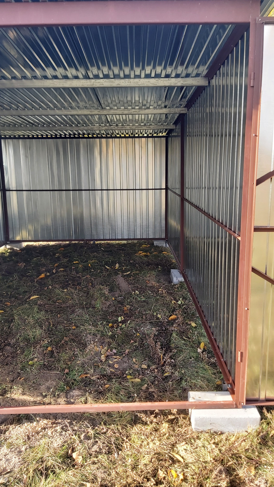 Wnętrze garażu blaszanego z widoczną konstrukcją szkieletową i blachą falistą. Widoczne betonowe bloczki fundamentowe i fragment podłoża z trawą i liśćmi.