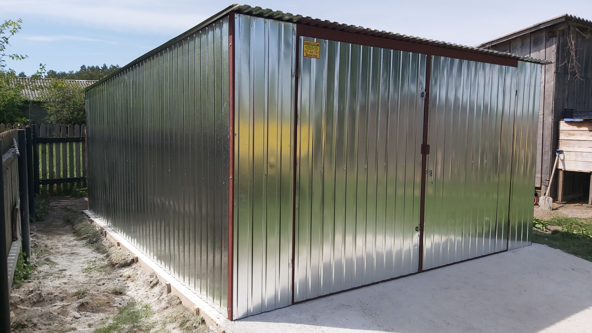 Nowy, blaszany garaż z dwuskrzydłowymi drzwiami, ustawiony na betonowej wylewce. Widoczne pionowe przetłoczenia blachy i brązowe obramowanie. Tło to zieleń i drewniane zabudowania.