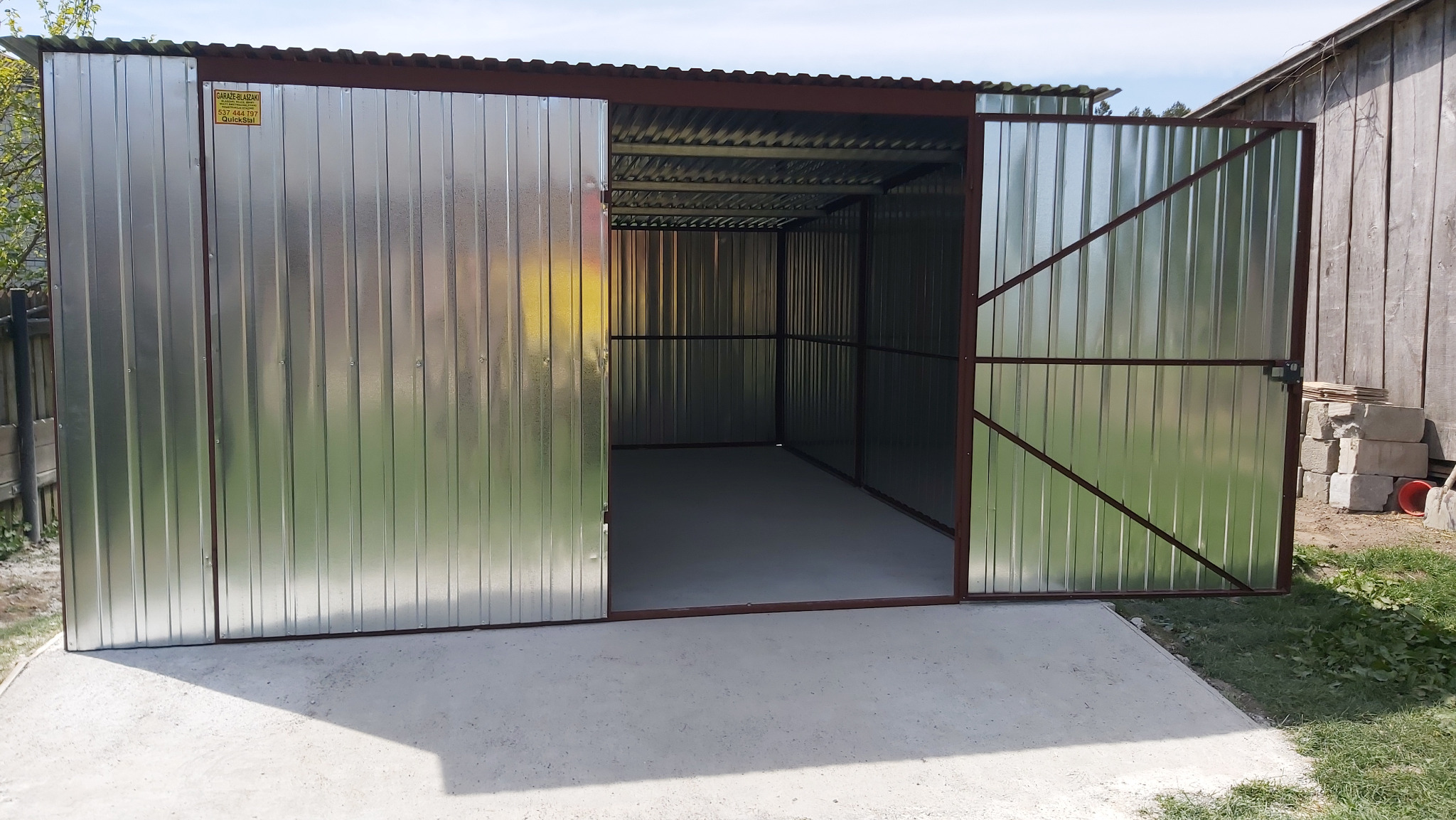 Nowy, blaszany garaż z otwartymi drzwiami na betonowej wylewce. Widoczna tabliczka informacyjna na ścianie. Garaż z brązową ramą i srebrnymi panelami.