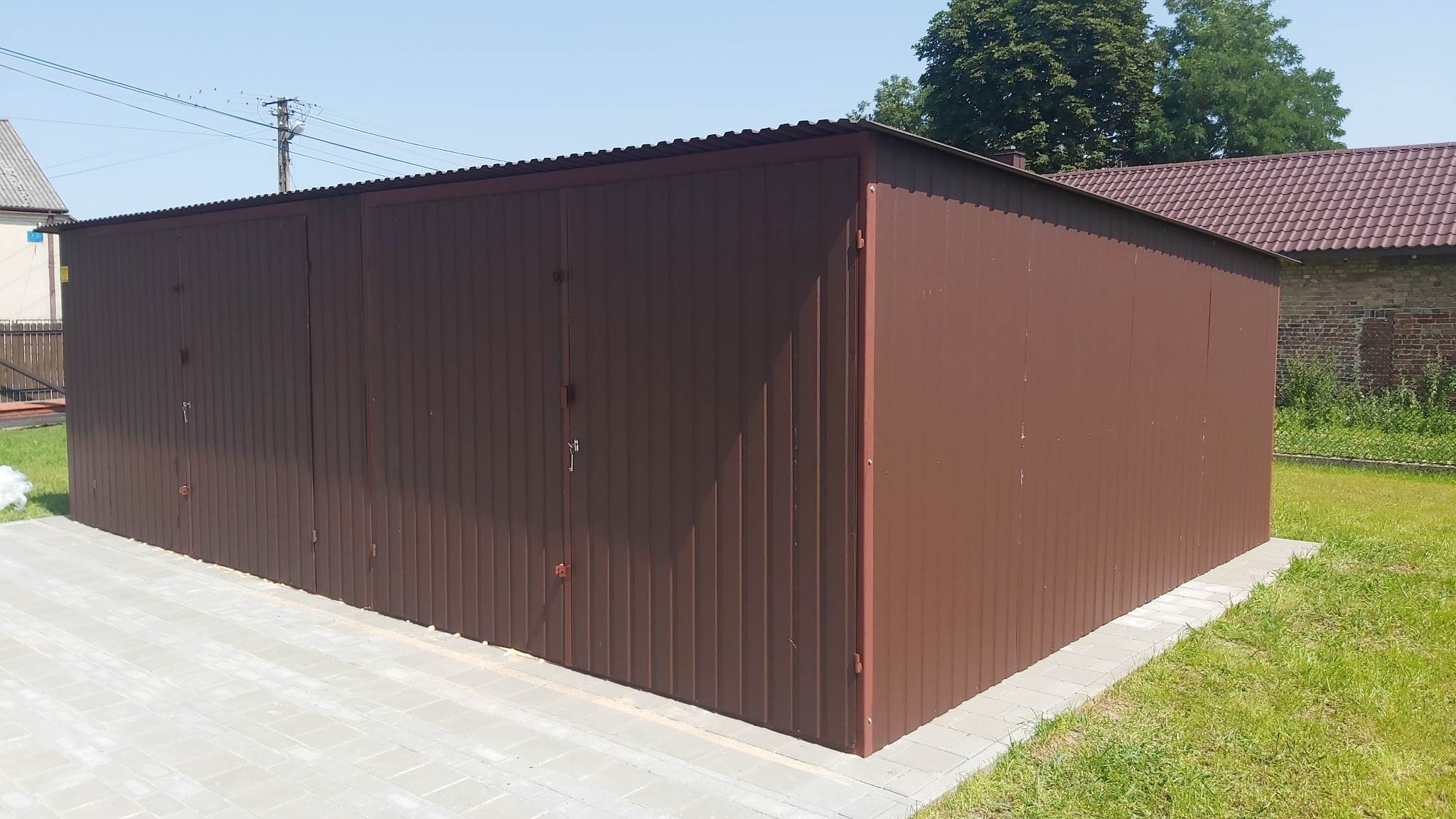 Brązowy garaż blaszany z dwoma parami drzwi, widoczny z perspektywy narożnika, na tle zielonego trawnika i ceglanego budynku.