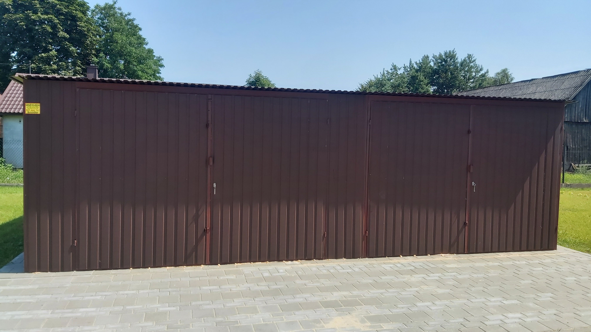 Brązowy garaż blaszany z dachem z blachy falistej na tle zieleni i nieba. Widoczne drzwi zamykane na klucz. Konstrukcja ustawiona na szarej kostce brukowej.