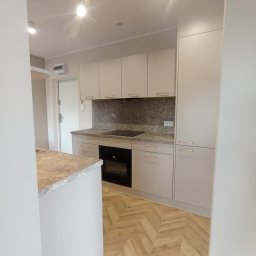 Meblo Complex - Jasna kuchnia z wyspą, fronty bez uchwytów, złote listwy, parkiet w jodełkę. Widok na zabudowę kuchenną z piekarnikiem i płytą. Minimalistyczny styl, nowoczesny design.