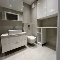 Nowoczesna łazienka z białymi meblami na wymiar: szafka pod umywalkę z szufladami, wisząca szafka z półkami. Szare płytki na ścianach i podłodze. Lustro z oświetleniem.