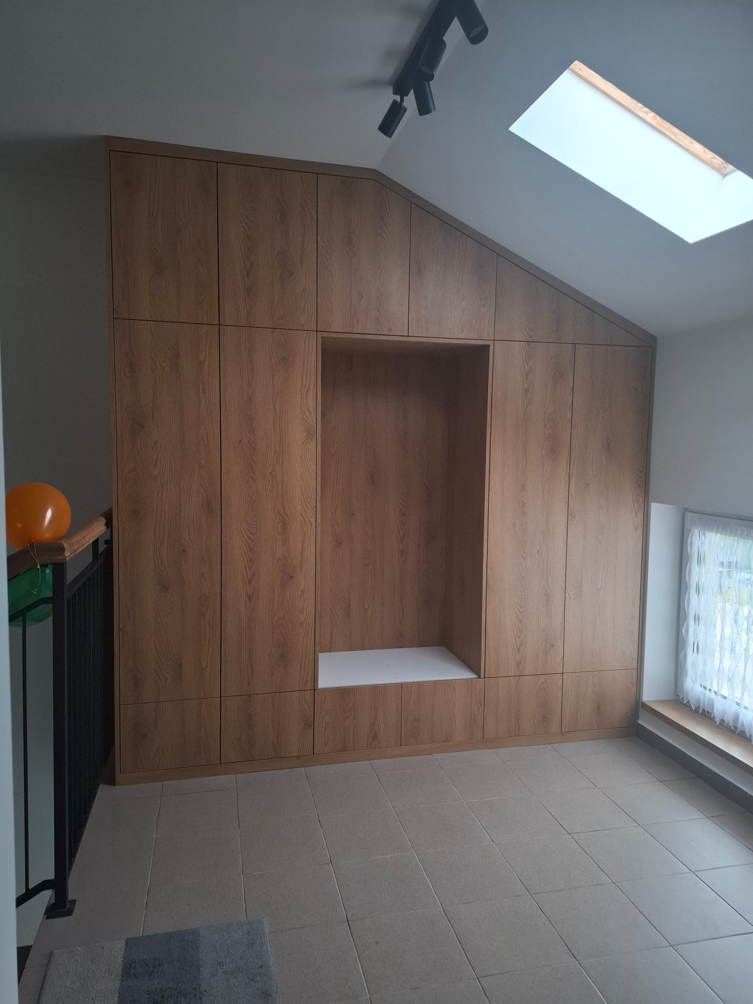 Drewniana szafa wnękowa z otwartą półką, dopasowana do skosu poddasza, z oknem dachowym w tle. Widoczne płytki podłogowe i fragment balustrady schodów.