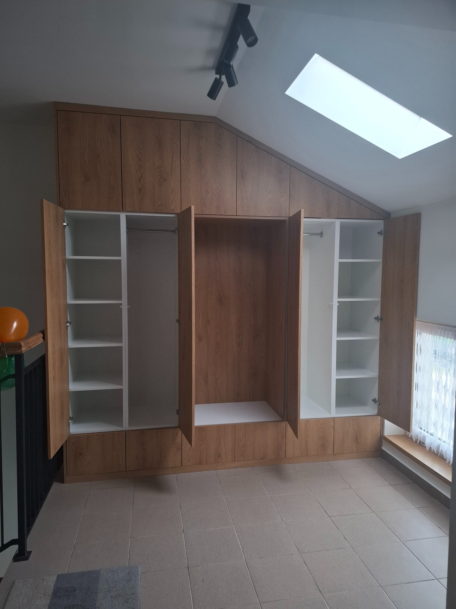 Szafa wnękowa z frontami w odcieniu drewna, dopasowana do skosu poddasza, z otwartymi drzwiami ukazującymi białe półki i drążki.