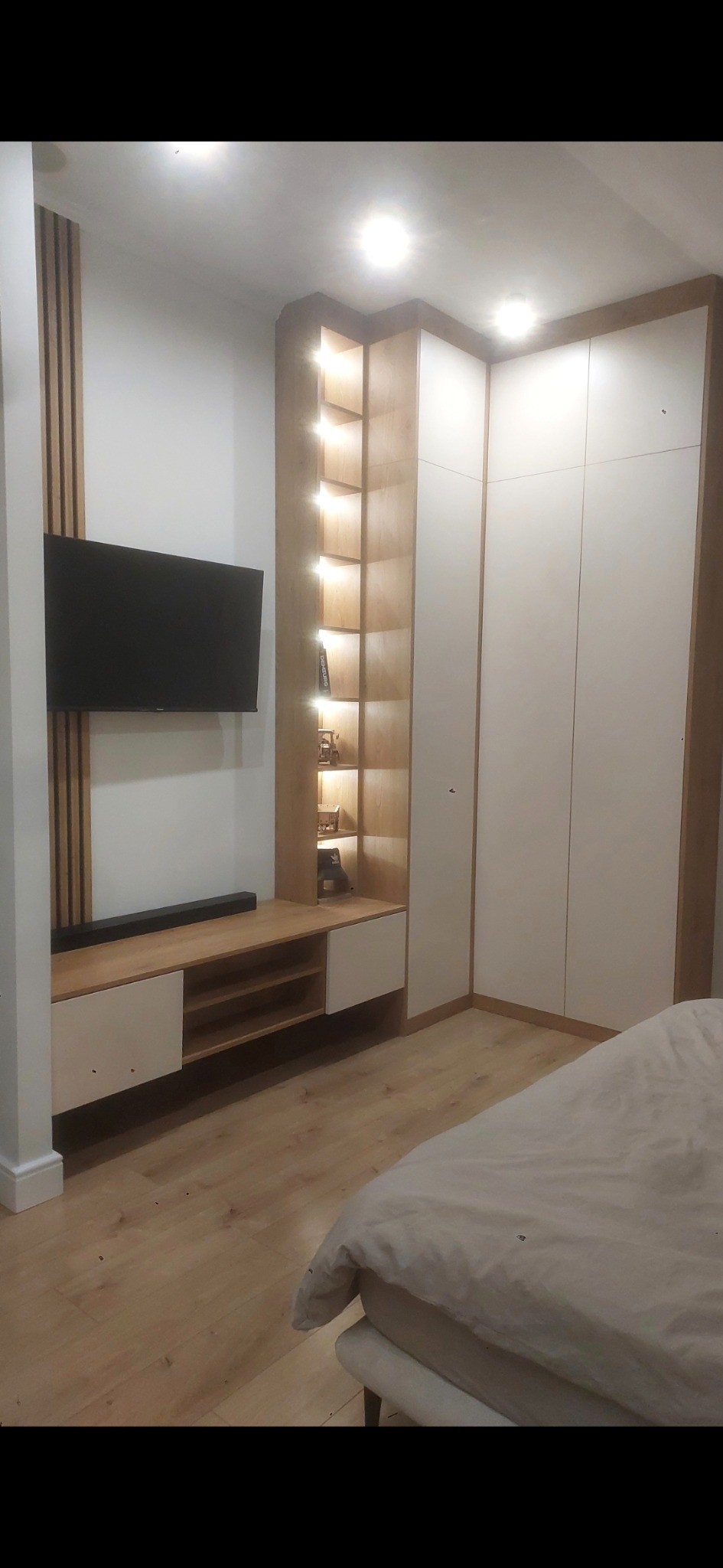 Biała szafa wnękowa z drewnianymi akcentami i półkami podświetlonymi LED, obok szafki RTV z czarnym telewizorem na ścianie. Nowoczesny design.