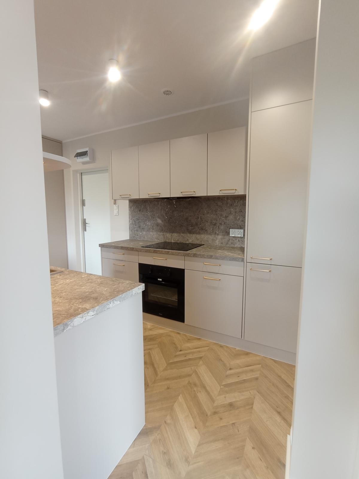 Jasna kuchnia z wyspą, fronty bez uchwytów, złote listwy, parkiet w jodełkę. Widok na zabudowę kuchenną z piekarnikiem i płytą. Minimalistyczny styl, nowoczesny design.