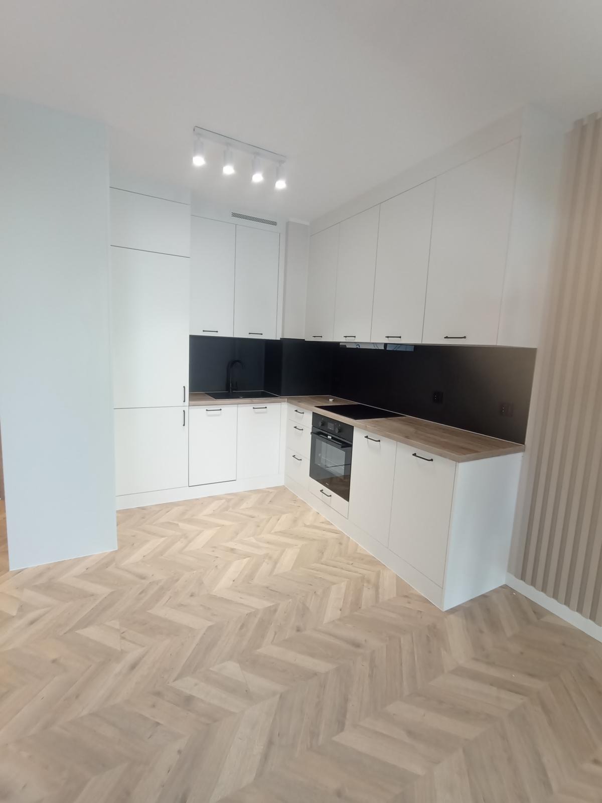 Jasna kuchnia z czarnym panelem nad blatem i drewnianym blatem. Parkiet w jodełkę. Minimalistyczne, podłużne uchwyty. Oświetlenie sufitowe punktowe.