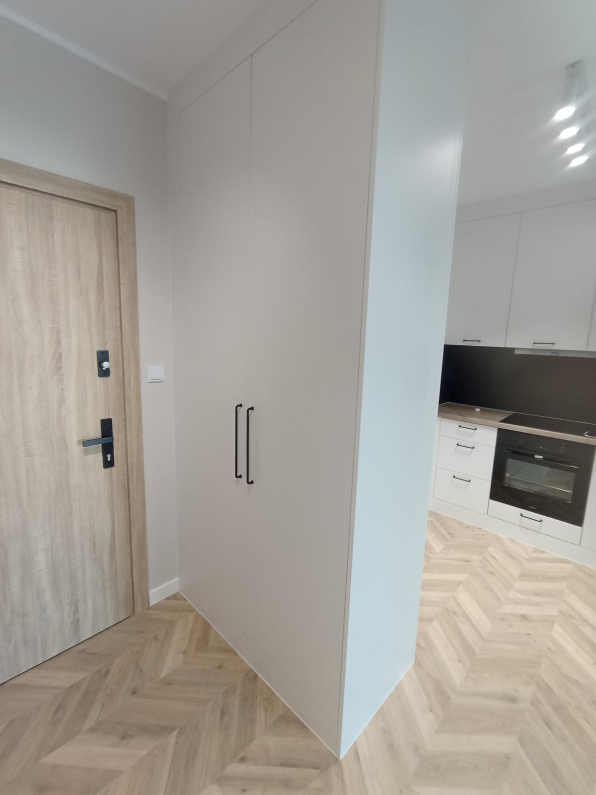 Biała szafa wnękowa z czarnymi uchwytami w przedpokoju, widok na jasną kuchnię z zabudową i sprzętem AGD, parkiet w jodełkę.