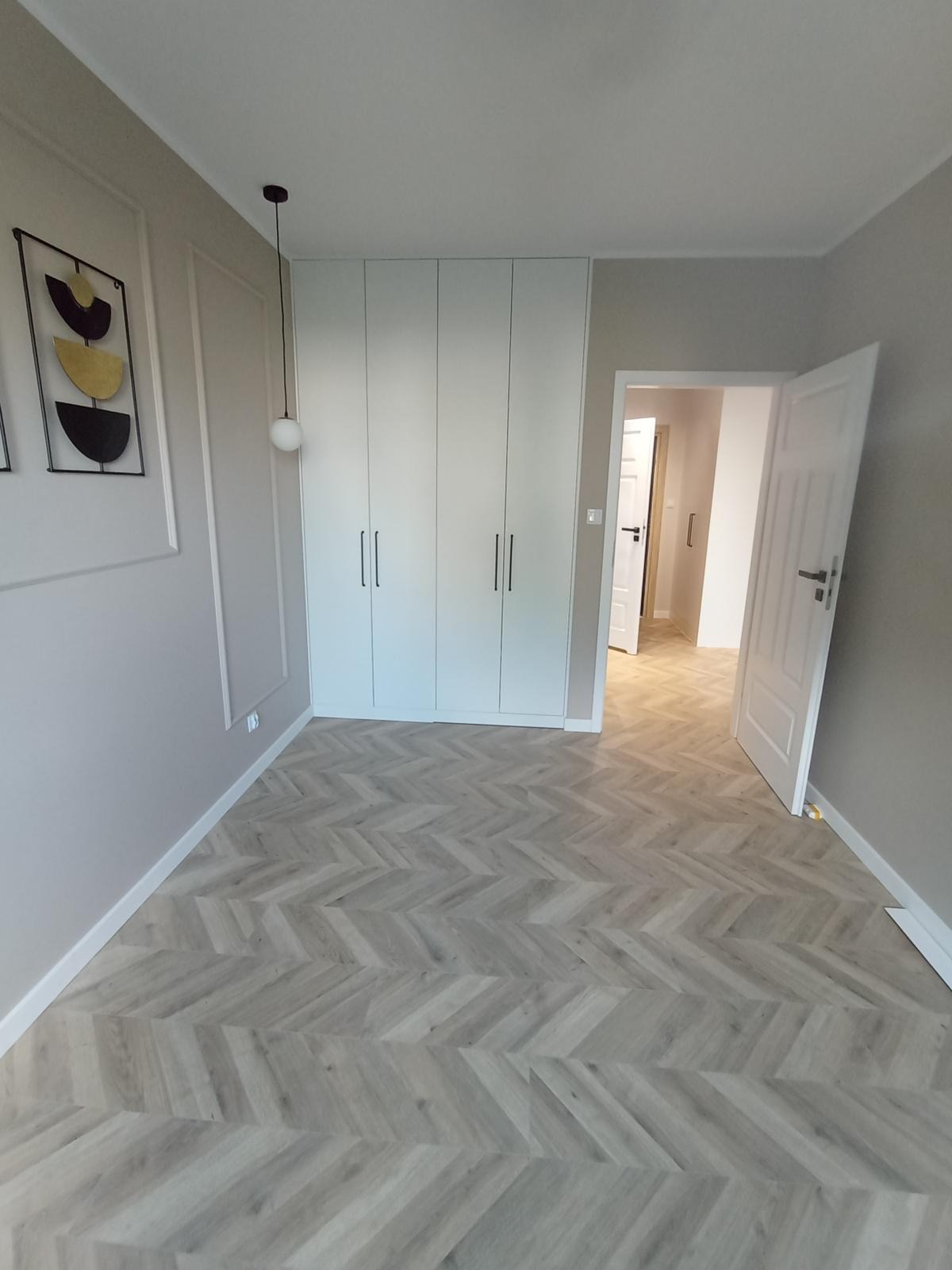 Jasne wnętrze z szafą wnękową pod sufit, parkietem w jodełkę i dekoracjami ściennymi. Otwarta przestrzeń z widokiem na dalsze pomieszczenia.