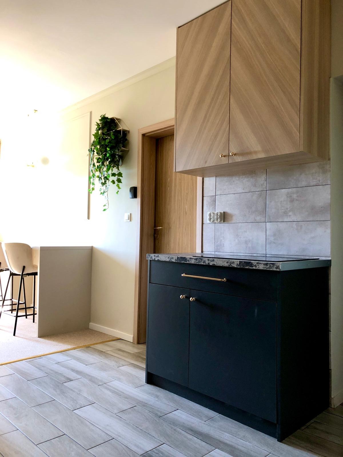 Ciemna szafka kuchenna z marmurowym blatem i złotymi uchwytami, nad nią szafka wisząca z drewnianymi frontami. W tle widoczna dekoracja z roślin.