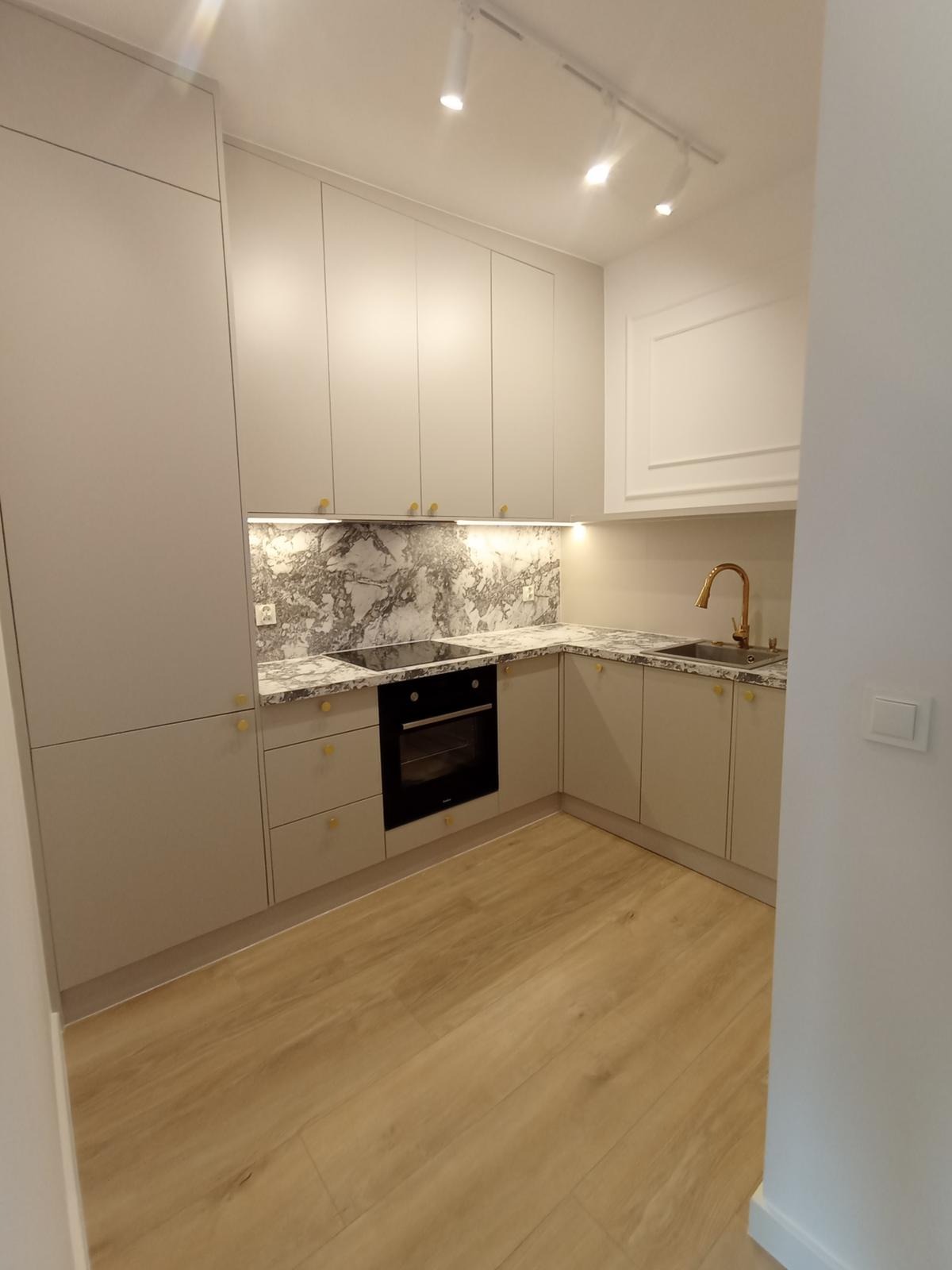 Jasna kuchnia na wymiar z marmurowym blatem i złotymi akcentami, oświetlona listwą LED pod szafkami, z drewnianą podłogą i nowoczesną armaturą.