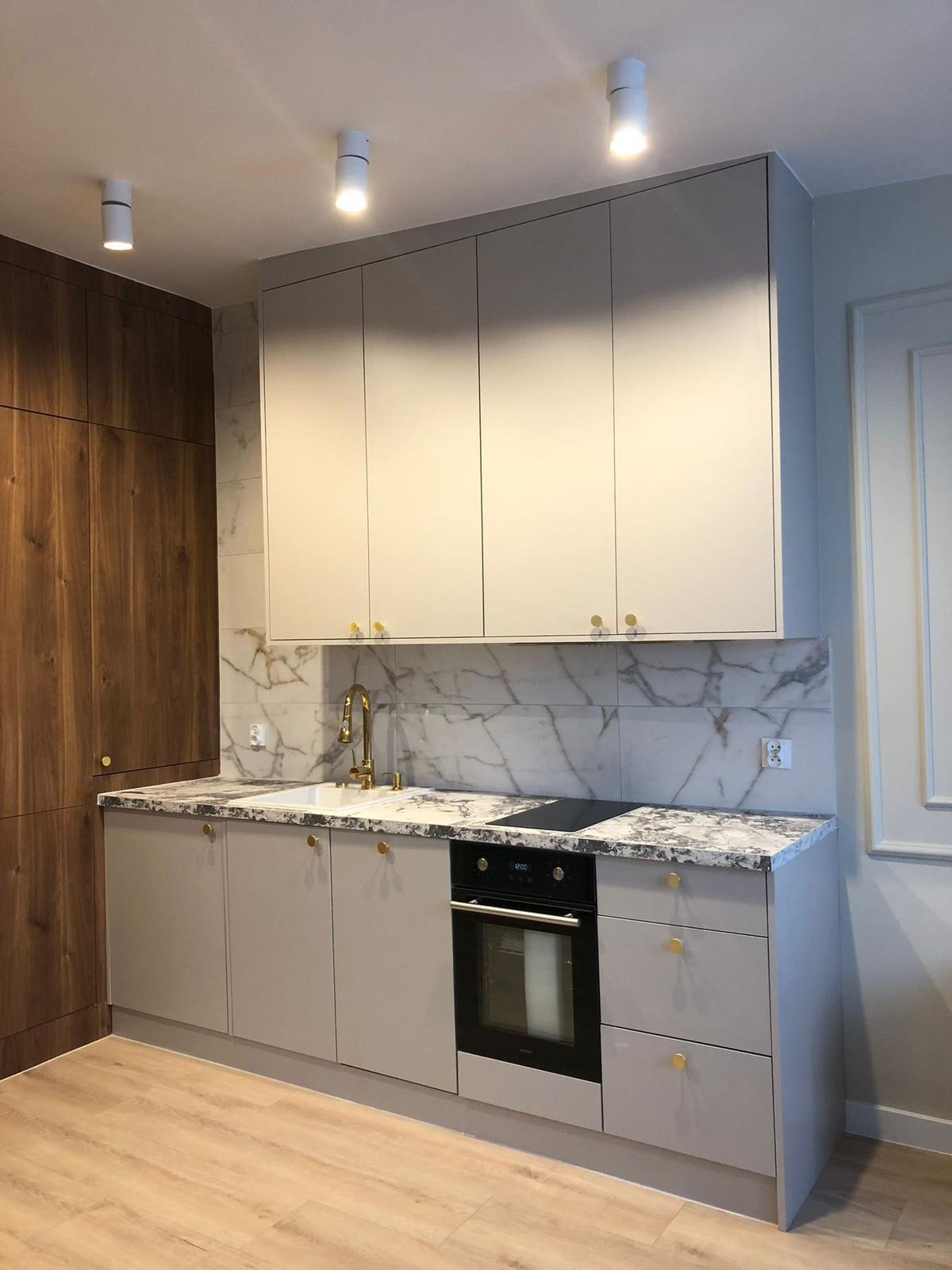 Jasna kuchnia z marmurowym blatem i złotymi uchwytami, widok na zabudowę z piekarnikiem i płytą indukcyjną, drewniana szafa po lewej stronie.