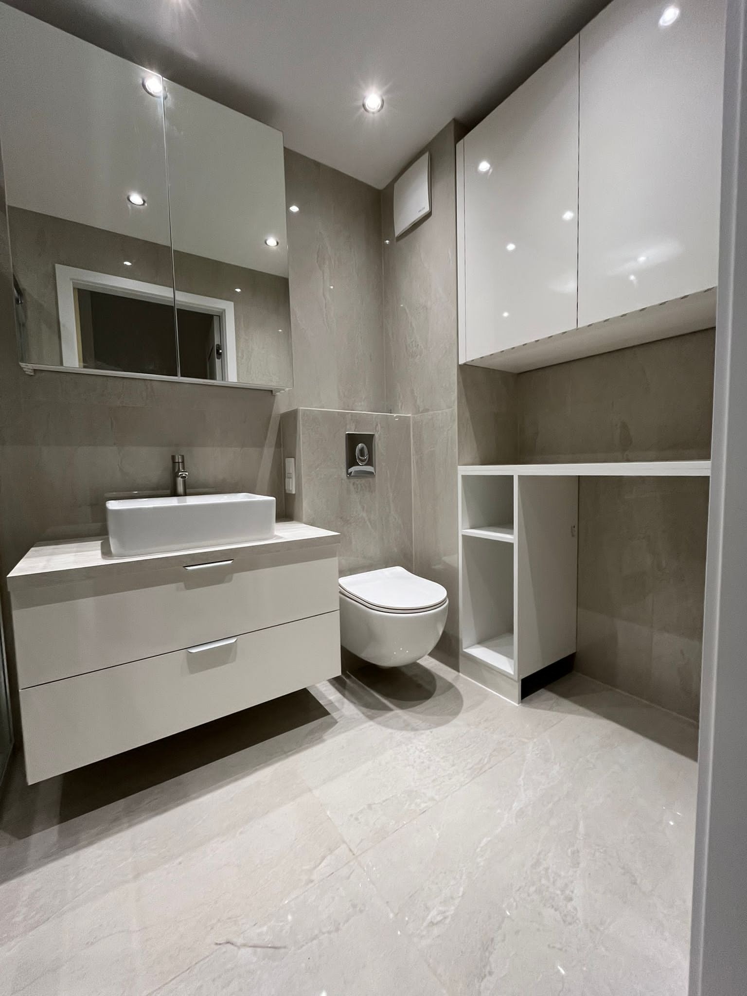 Nowoczesna łazienka z białymi meblami na wymiar: szafka pod umywalkę z szufladami, wisząca szafka z półkami. Szare płytki na ścianach i podłodze. Lustro z oświetleniem.