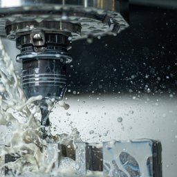 ZOK-TECH - Precyzyjna obróbka metalu na maszynie CNC z chłodziwem, w zbliżeniu. Wióry i płyn chłodzący w ruchu, metalowy element obrabiany. Szare tło.