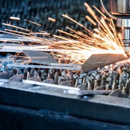 ZOK-TECH - Precyzyjne cięcie metalu laserem, iskry w locie, fragmenty obrobionego materiału na perforowanym stole. Detal maszyny w tle, chłodna kolorystyka.