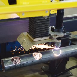 ZOK-TECH - Automatyczne cięcie plazmowe rury stalowej z trzema wyciętymi otworami. Iskry i jasne światło w miejscu cięcia. Żółta rama maszyny. Obróbka CNC.