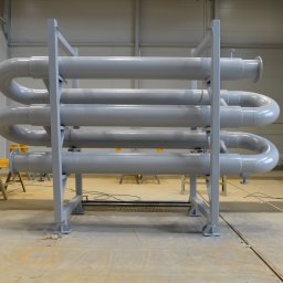 ZOK-TECH - Szara, stalowa konstrukcja z rurami ułożonymi w spiralę, na tle hali produkcyjnej. Widoczne elementy wsporcze i podesty malarskie. Charakterystyczne połączenia kołnierzowe.