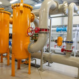 ZOK-TECH - Instalacja przemysłowa z żółtymi zbiornikami i szarymi rurami, widoczne zawory z czerwoną armaturą. Jasne oświetlenie hali, białe ściany i podłoga.